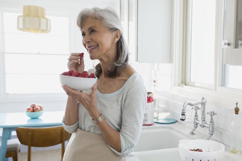 An older woman eats raspberries from a bowl in her kitchen.