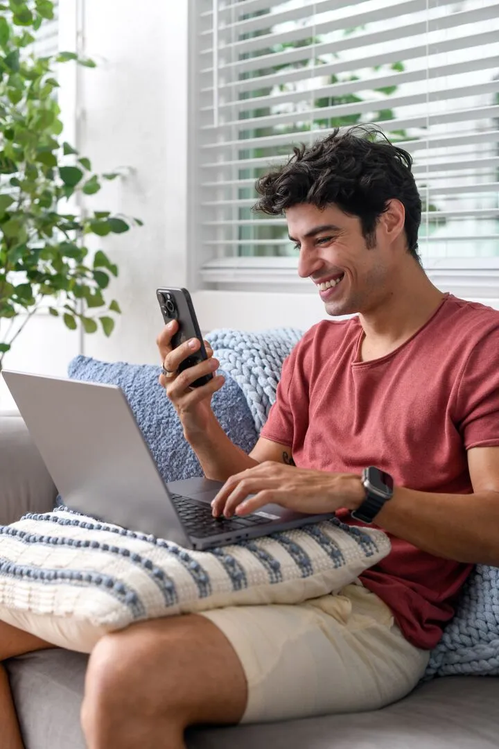 Man sitting on sofa looking at App on phone with computer open
